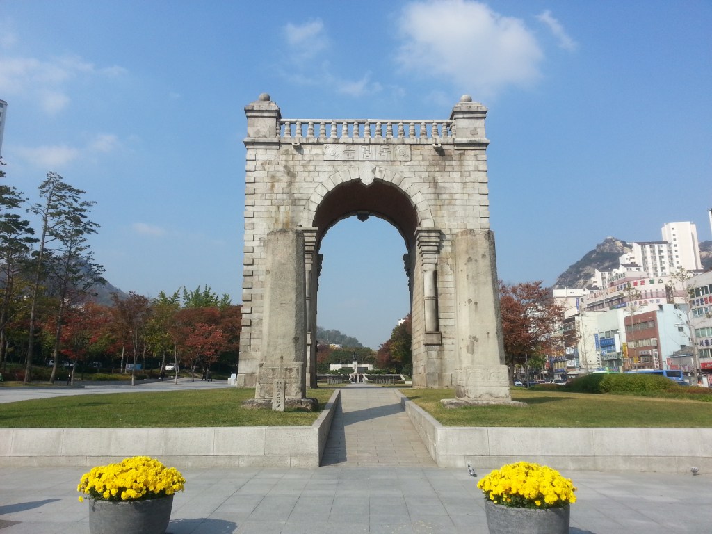 Independence Gate, a historic monument in Seoul, stands tall with a clear blue sky above and vibrant autumn foliage in the background. Yellow chrysanthemums are planted in stone pots in the foreground, adding color to the serene park setting.