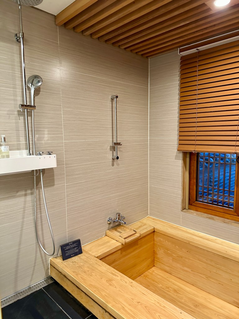 A wooden bathtub with a shower area, featuring wooden accents and a window with traditional Korean lattice, in a modern bathroom setting.
