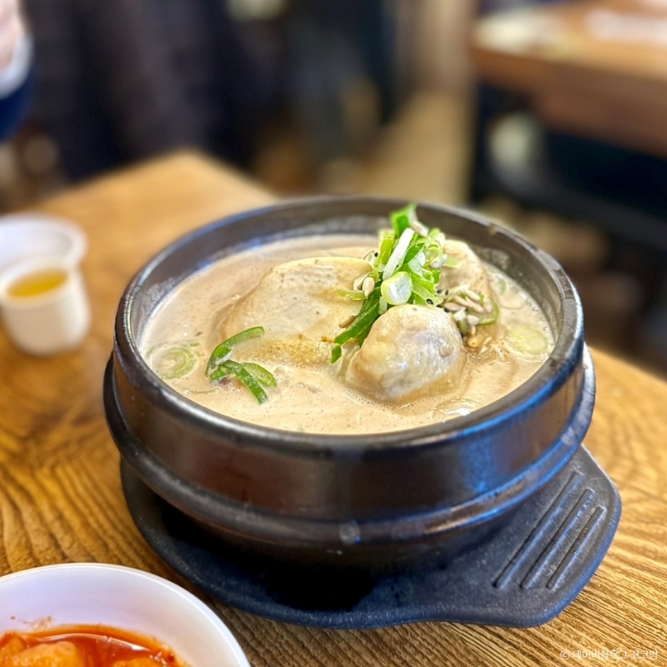 A bowl of ginseng chicken soup (samgyetang) served in a traditional stone pot, garnished with green onions, accompanied by a side of kimchi.